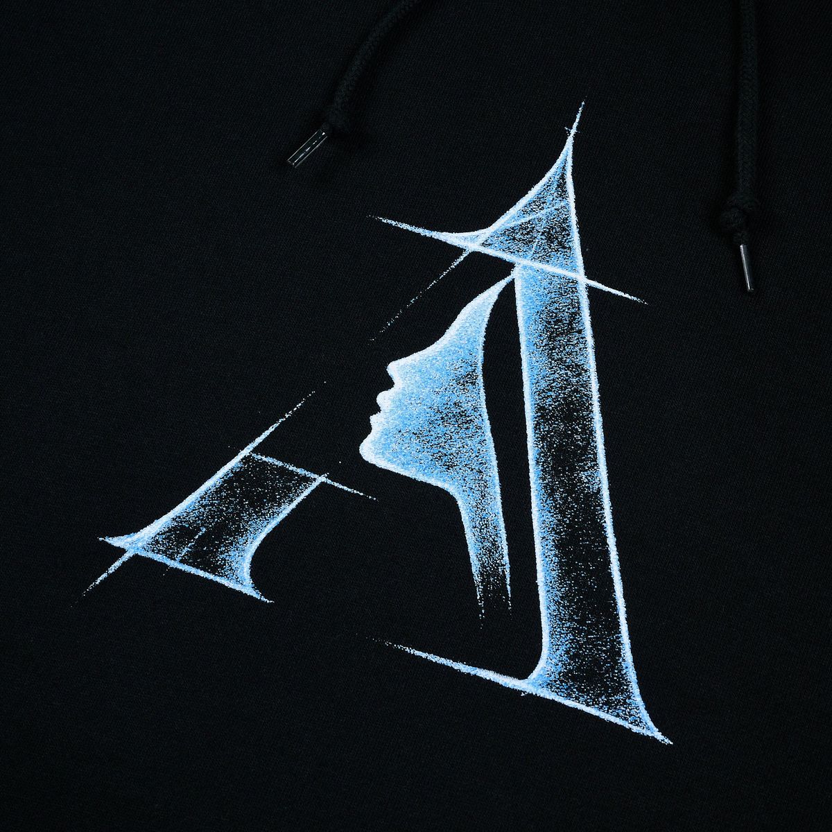 Close-up of a black april skateboards hoodie featuring a stylized graphic print of the letter 'A' in a light blue, textured, speckled dot pattern. The hood drawstrings are visible at the top.