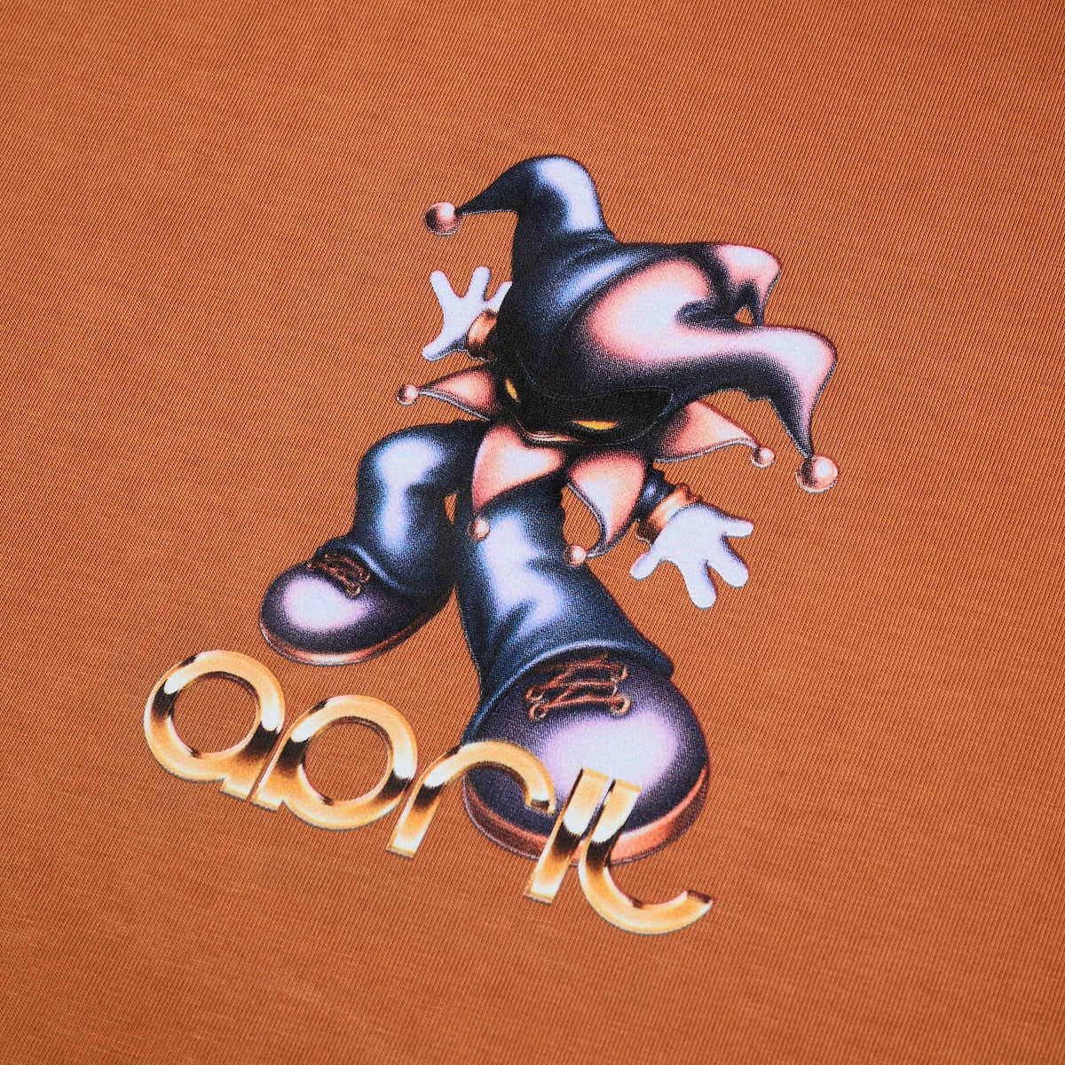 A close-up of a brown t-shirt featuring a graphic print of a jester in black and pink with white gloves, above the word 'April' in metallic gold script.