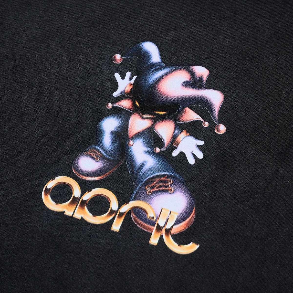 Close-up of a black April skatebaords t-shirt featuring a graphic of a stylized jester in dark blue and pink with white gloves, above the word 'april' in metallic gold 3D lettering.